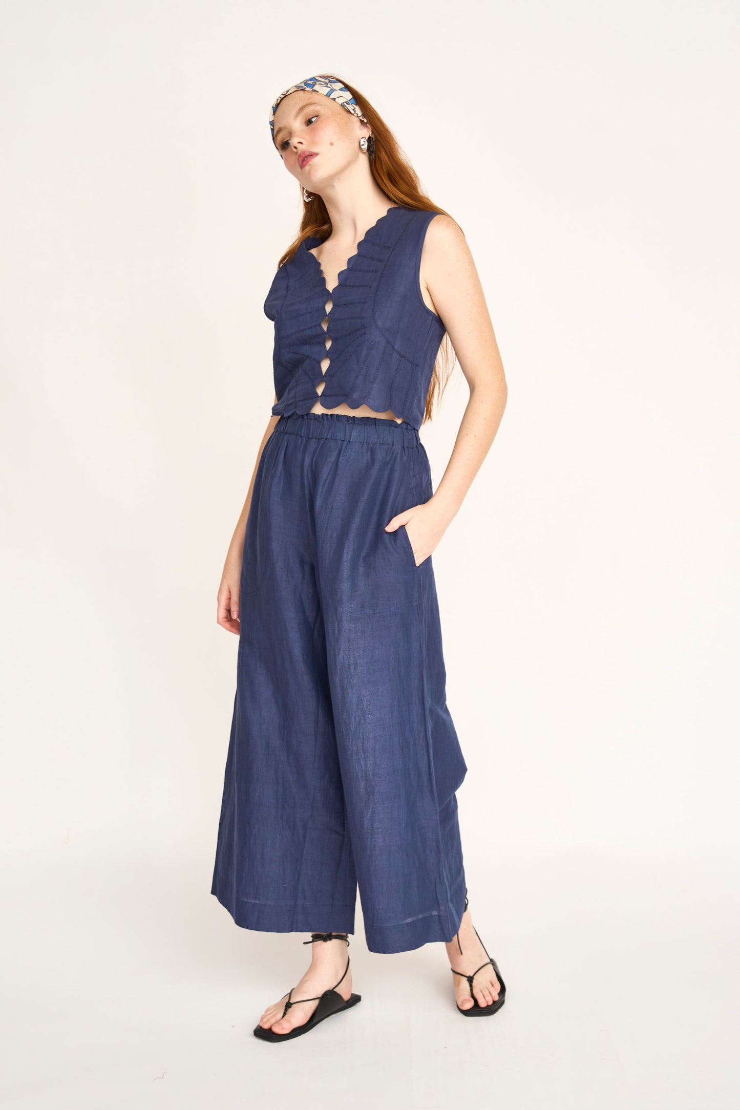 Woman in a blue cotton top, wide-leg pants, headband, and sandals stands against a plain background.