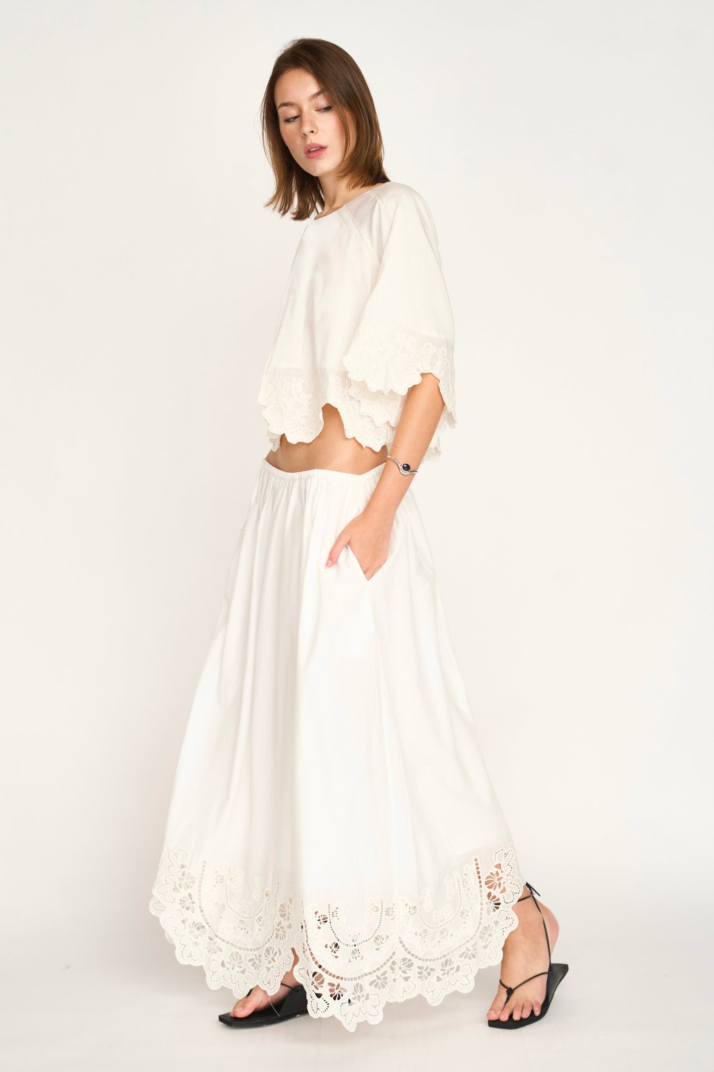 Woman in a white eyelet crop top and long embroidered white skirt, shown against a plain background.