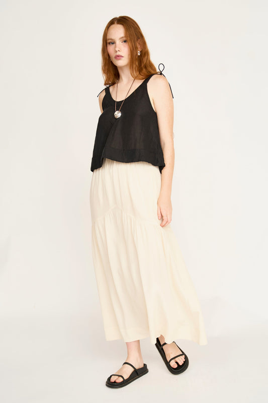 Cream silk skirt, black sleeveless top, sandals, and necklace worn by a woman standing against a white background.
