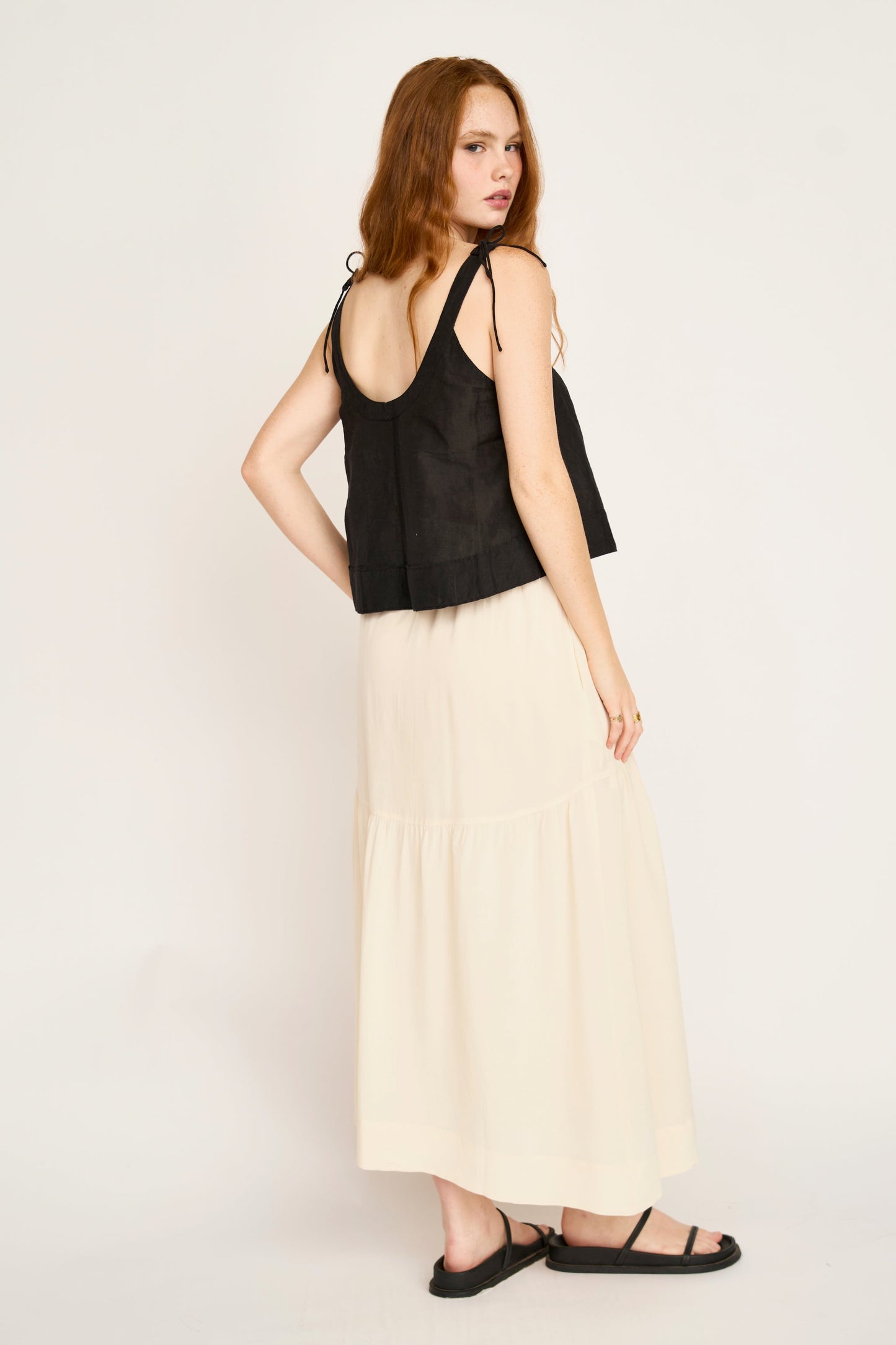 Woman with long red hair in a cream silk skirt, black sleeveless top, and black sandals.