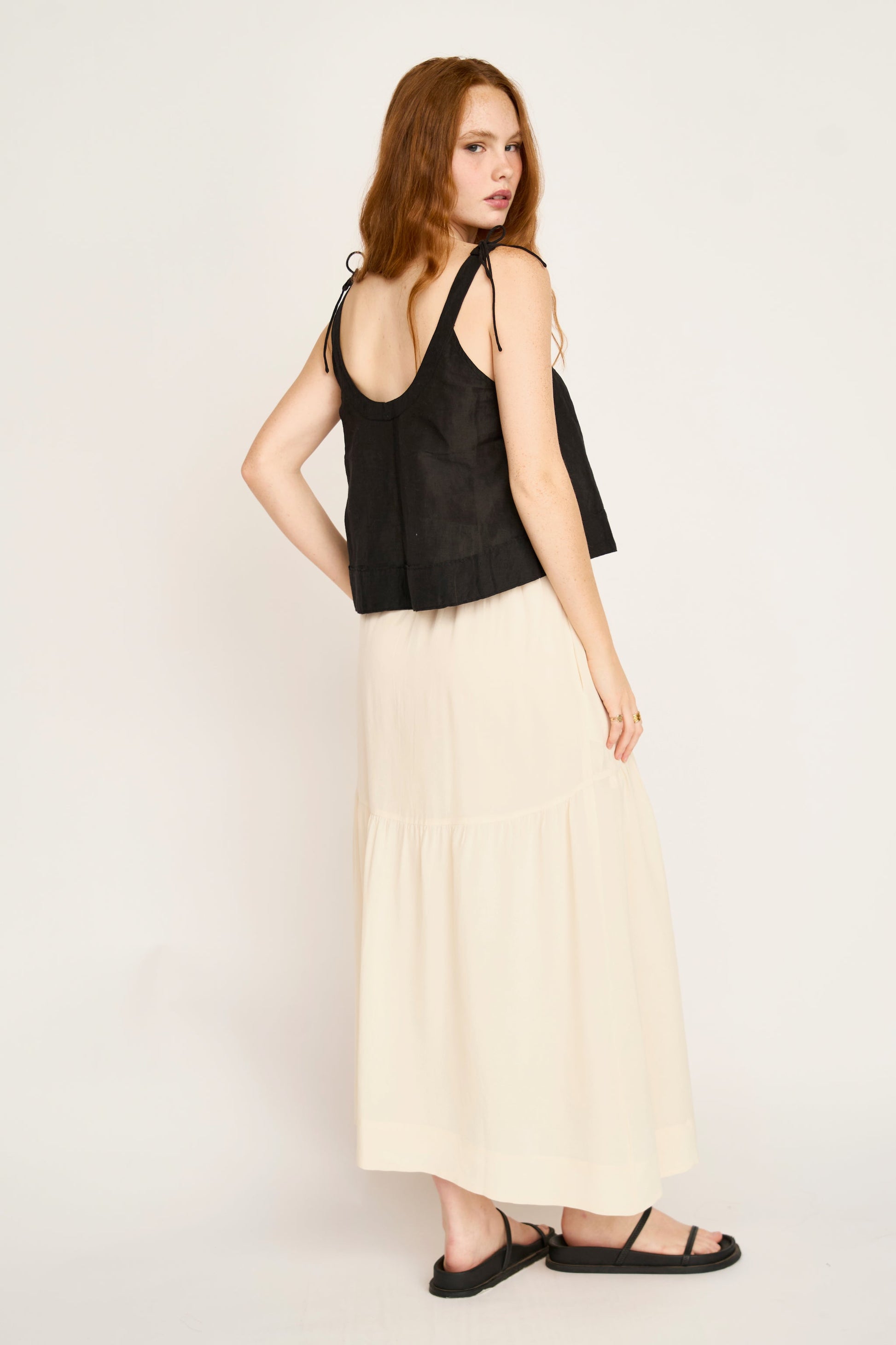 Woman with long red hair in a cream silk skirt, black sleeveless top, and black sandals.