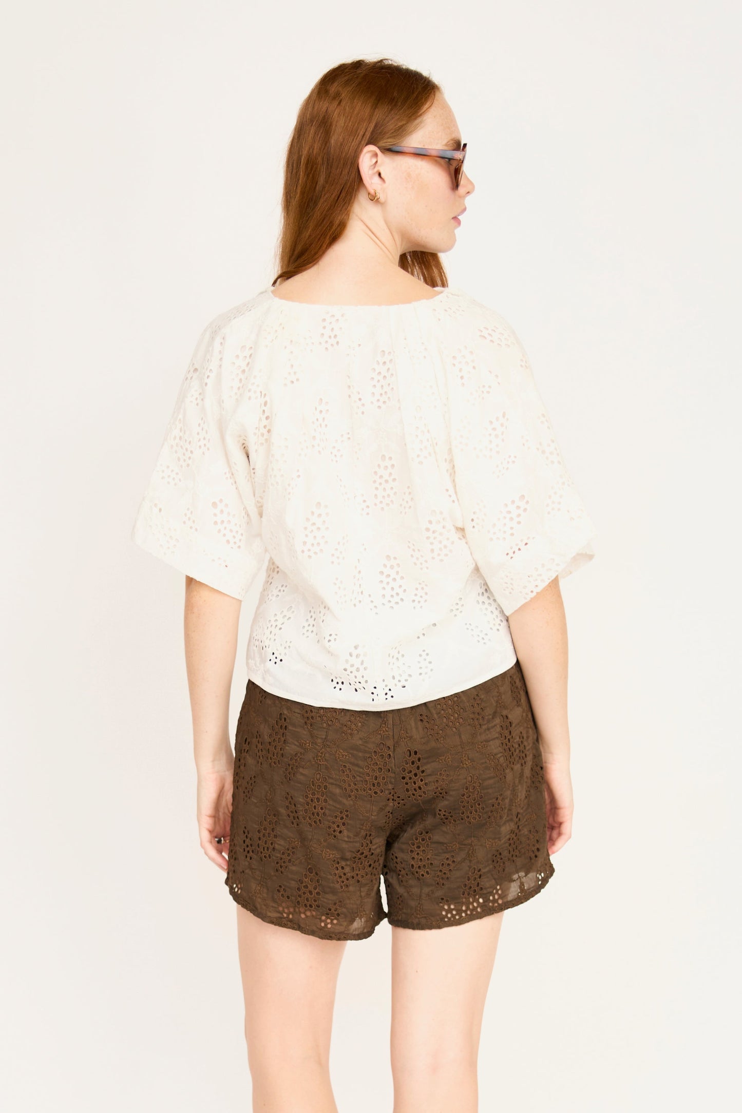 Woman in sunglasses wears a white eyelet cotton top with brown shorts, shown from the back.