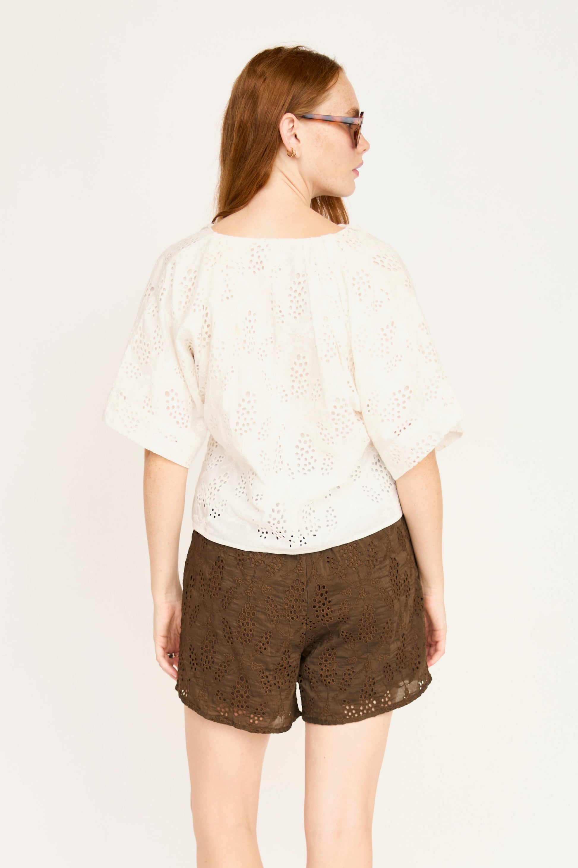 Woman in sunglasses wears a white eyelet cotton top with brown shorts, shown from the back.
