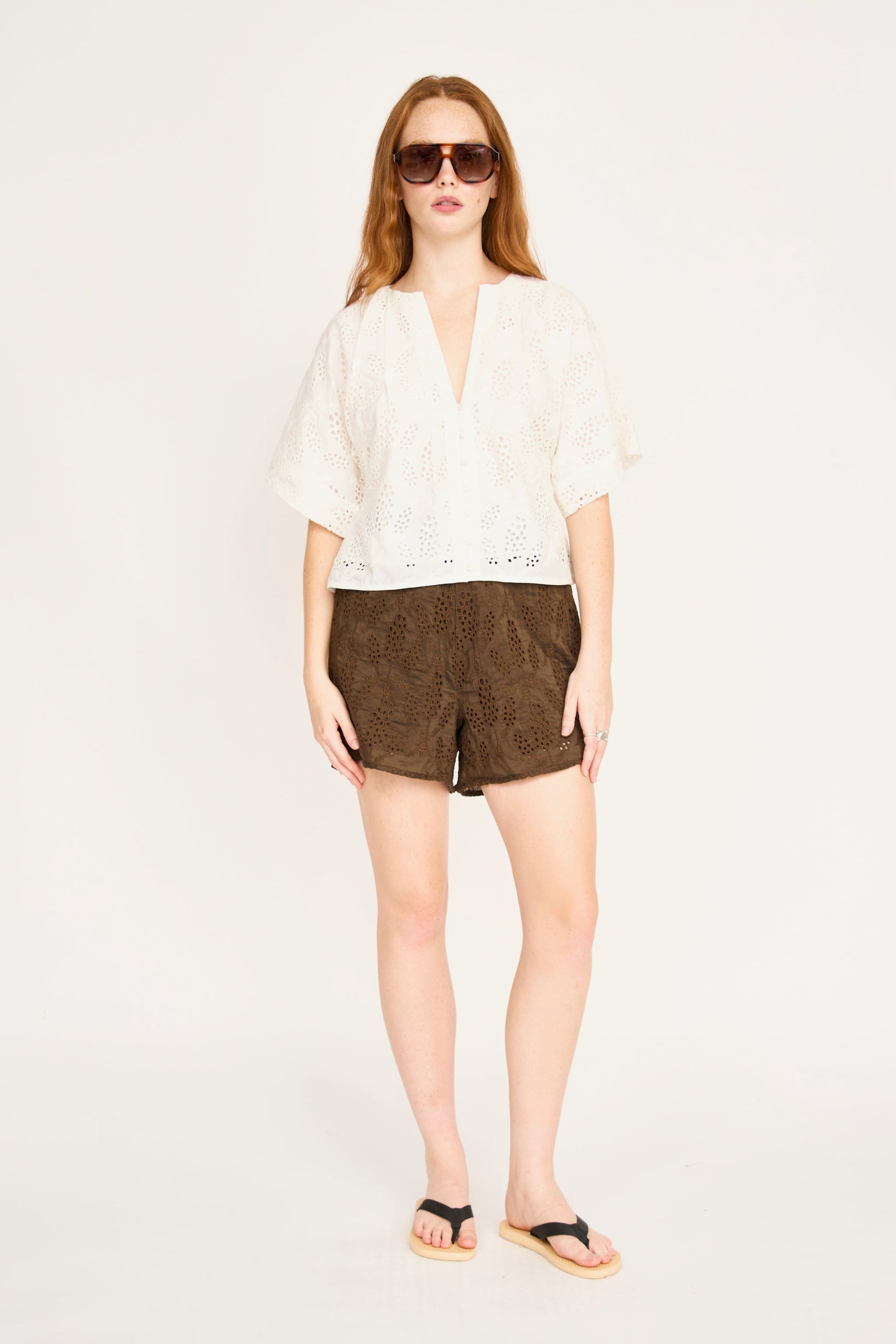 Woman in sunglasses wearing white eyelet top, brown shorts, and flip-flops.