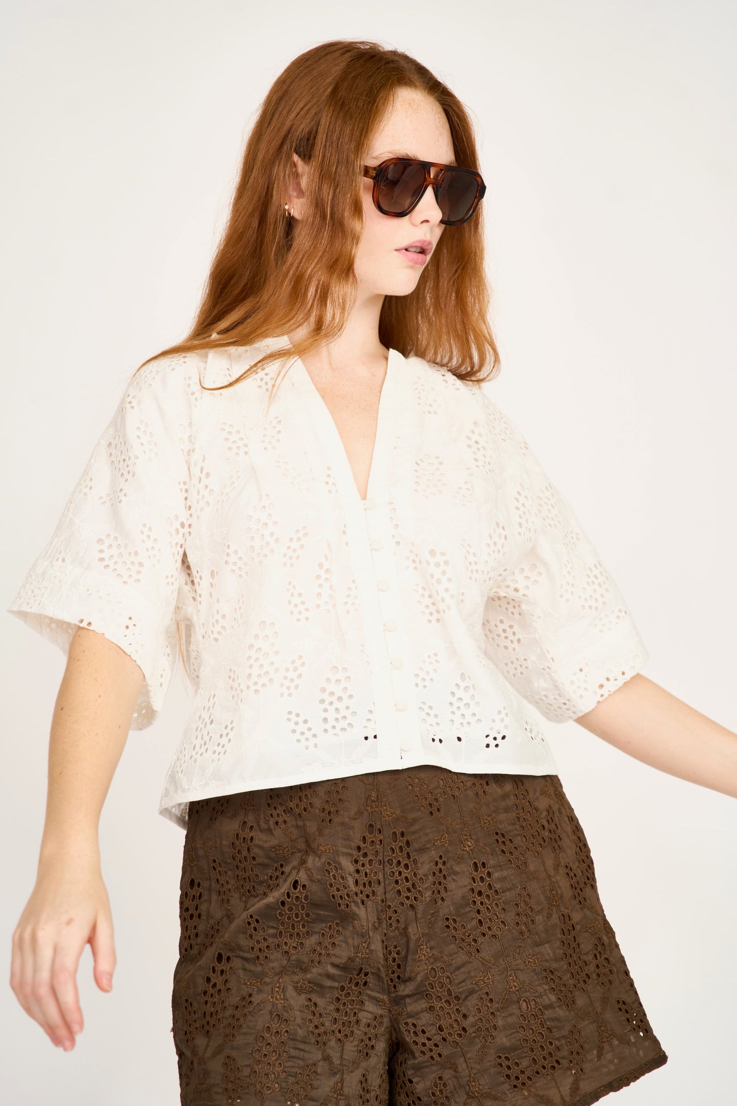 Woman in sunglasses models a white eyelet blouse with long sleeves against a plain background.