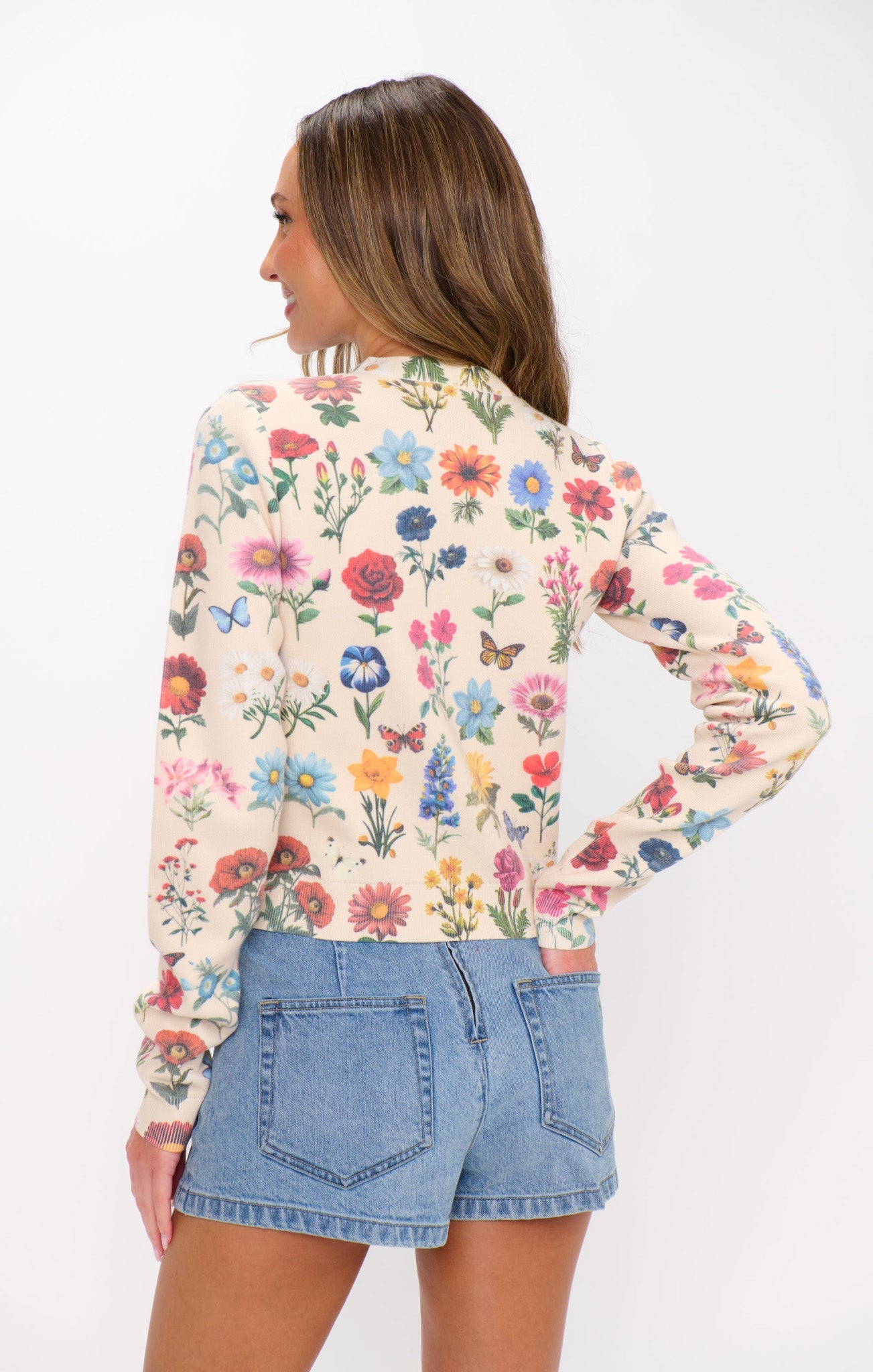 Back view of a woman in a floral knit cardigan and denim shorts, standing against a plain white background.