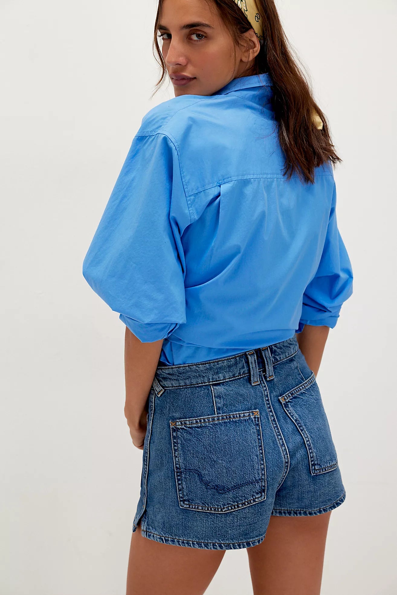 Woman in a blue shirt and dark denim sailor shorts, facing away, head turned, hair secured with a scarf.