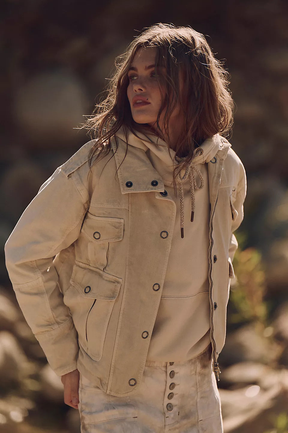 Woman wearing a light beige washed cotton jacket stands outside, gazing sideways with tousled hair.