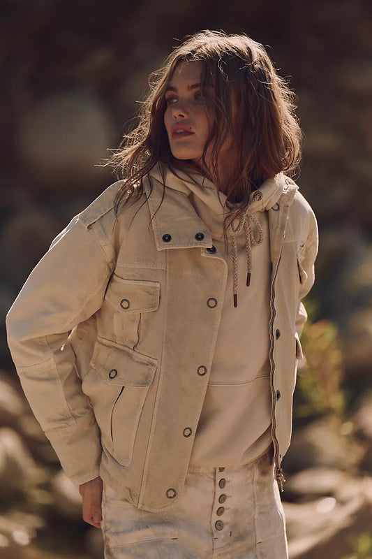 Woman wearing a light beige washed cotton jacket stands outside, gazing sideways with tousled hair.