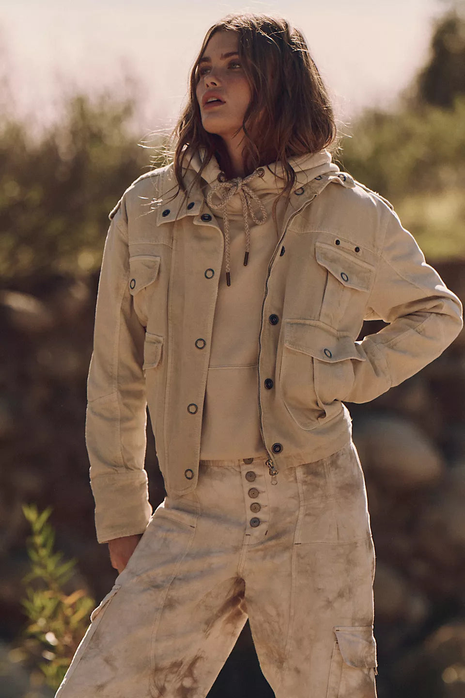 Woman outside wearing a tan washed cotton jacket over a light hoodie and tie-dye pants, looking into the distance.
