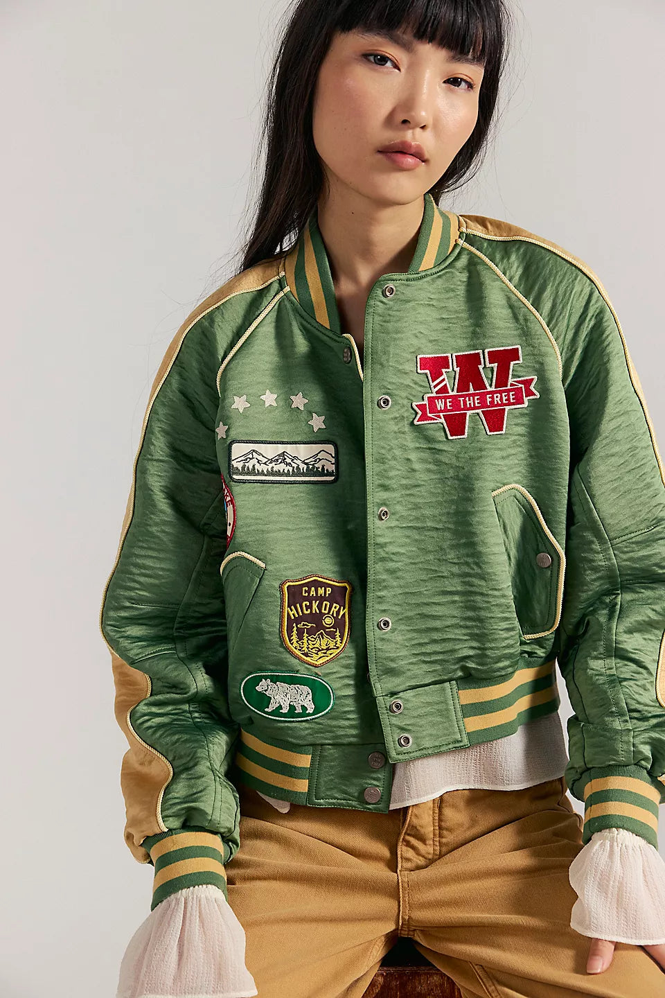 Woman wearing a vintage green souvenir jacket and tan pants sits on a stool. Jacket features embroidered detailing.