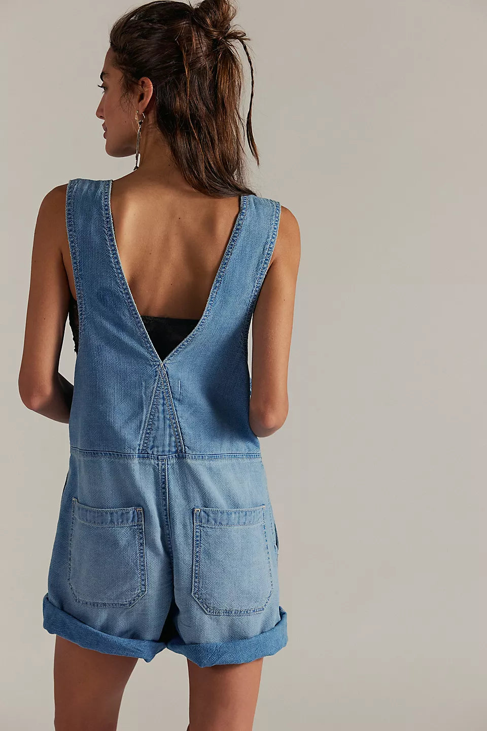 Woman with long hair in light indigo denim shortalls with a deep V-back and rolled shorts, suitable for casual wear.