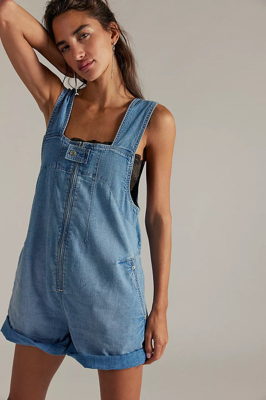 Woman in light indigo denim shortall over a black top, one arm raised behind her head.