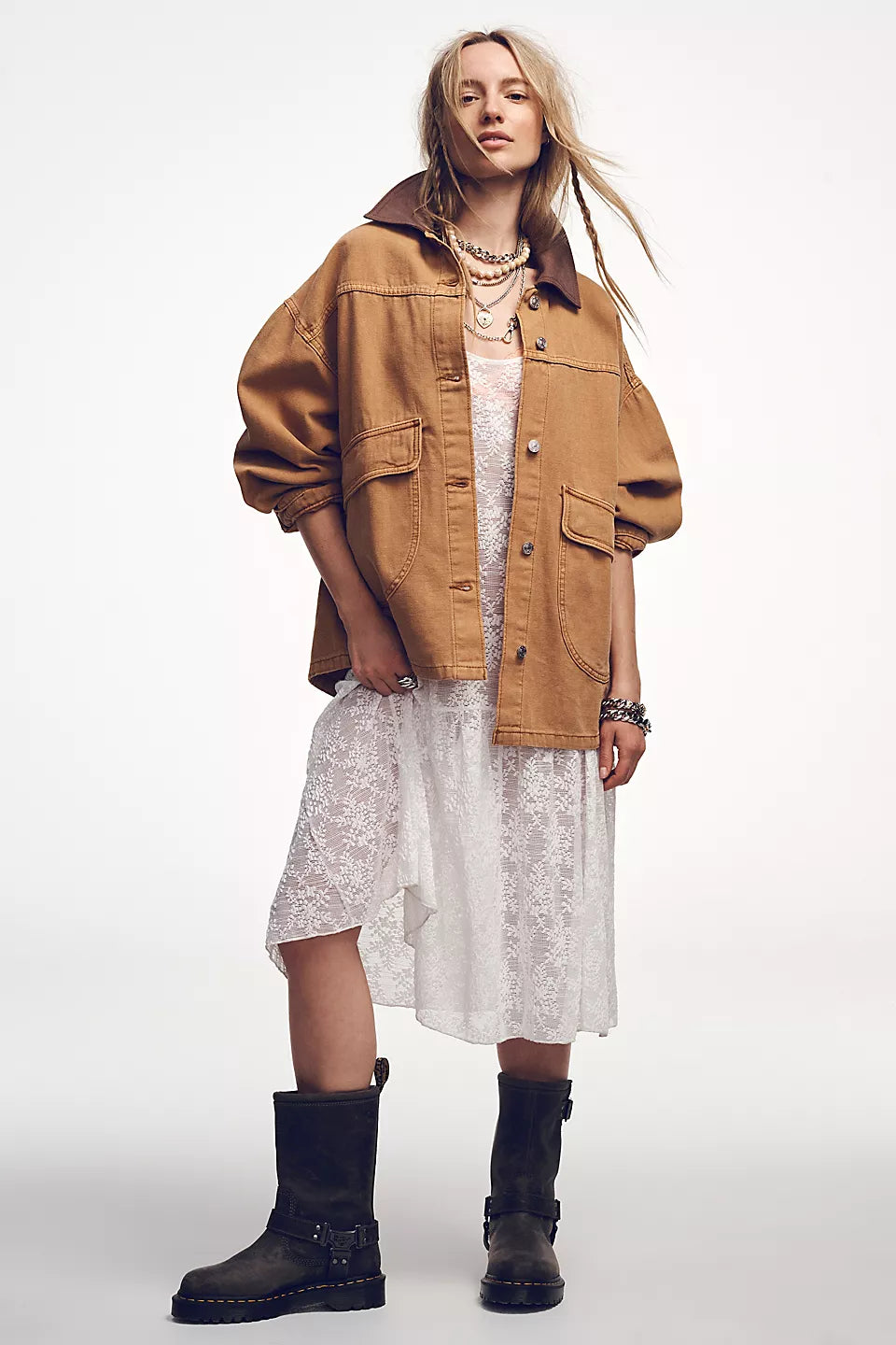 Woman wears a white lace dress, tan denim jacket, layered metal necklaces, and black leather boots.