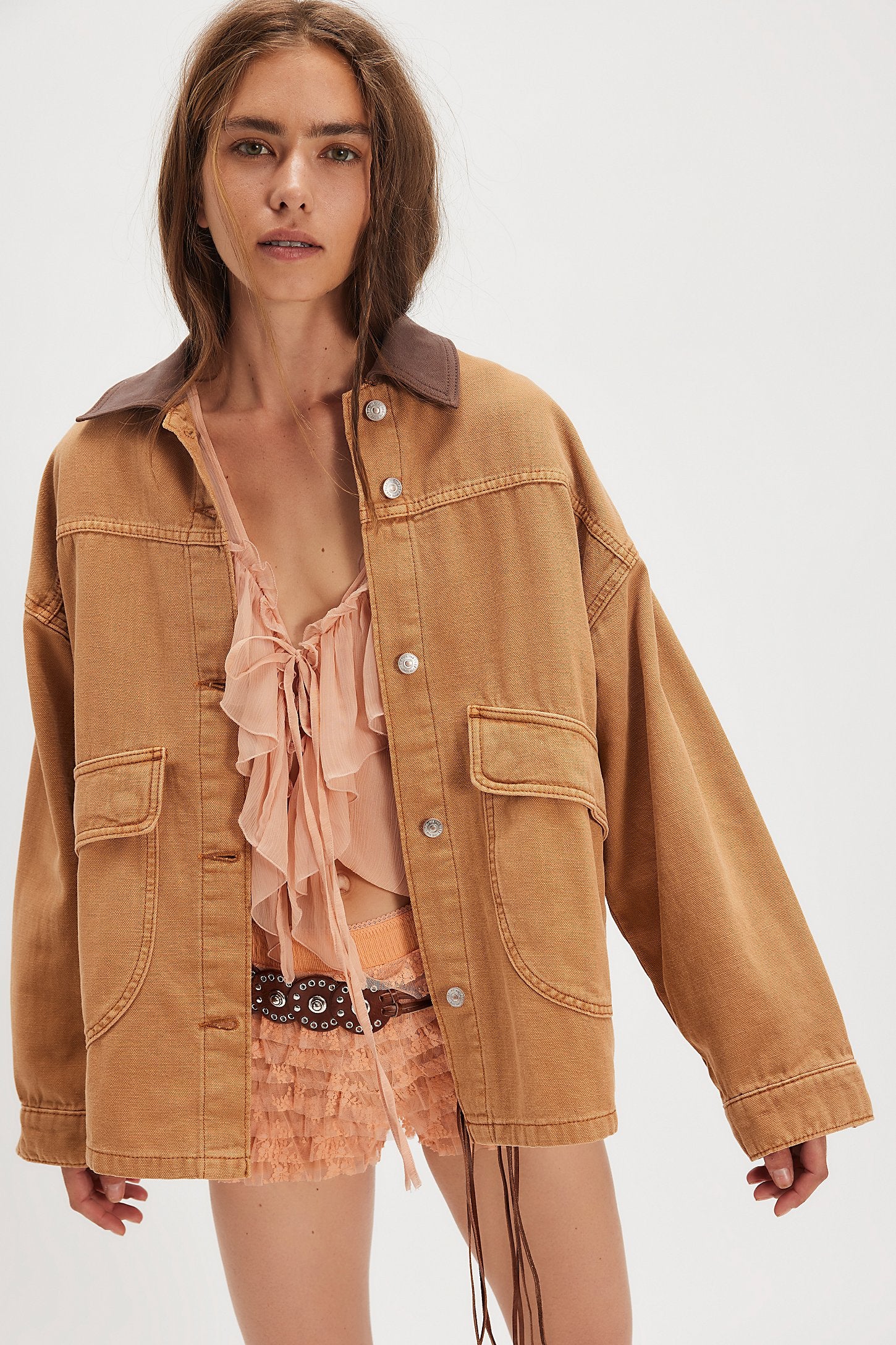 Woman in a tan denim jacket over a peach ruffled top and shorts stands against a plain backdrop.