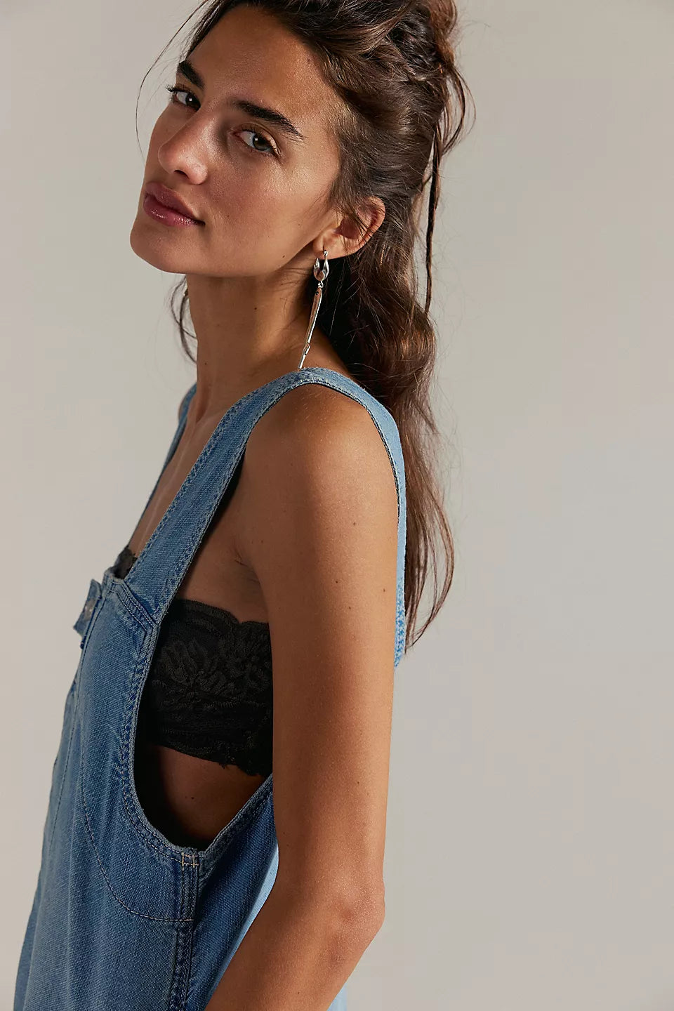 Woman with long brown hair models a light indigo denim shortall over a black lace bralette, gazing softly.