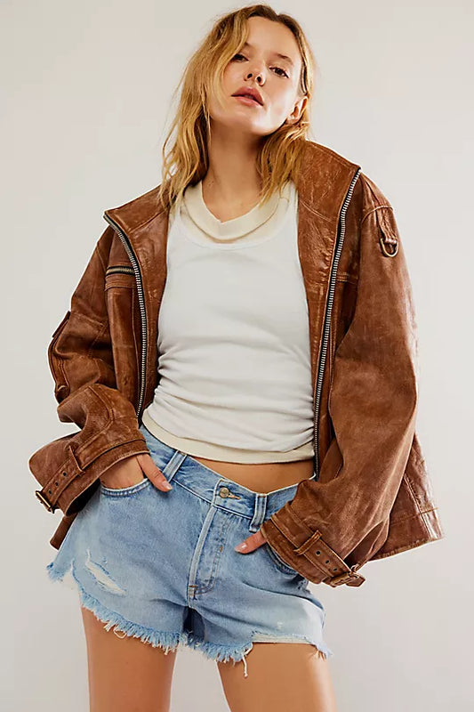 Woman in denim shorts, brown leather jacket, and white top stands against a plain background.