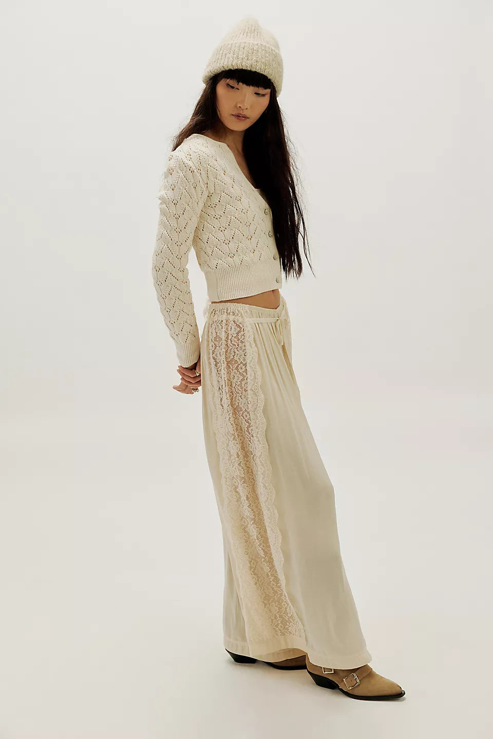 Woman in ivory knit cardigan, long lace skirt, tan boots, and a beanie poses indoors.
