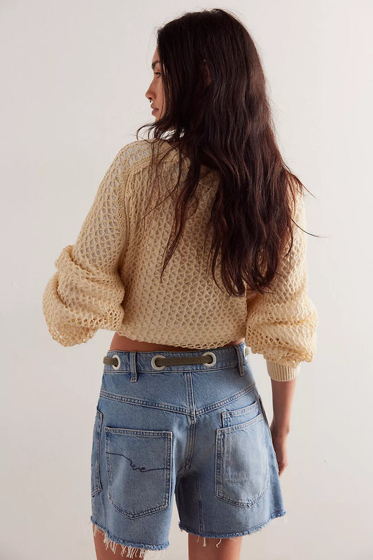 Woman with long dark hair wears a beige knit sweater and blue shorts, facing away to show the back detail of both garments.