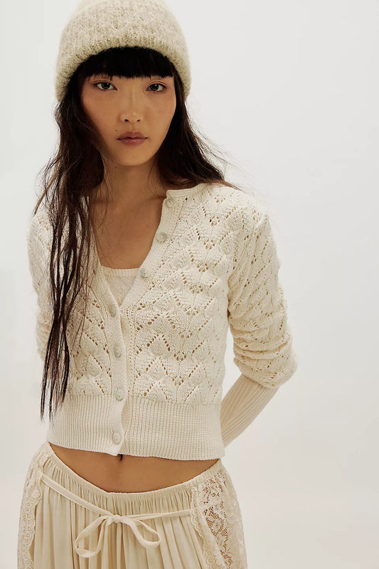 Woman wearing an ivory pointelle cardigan, matching knit top, lace-trim pants, and a cream beanie.