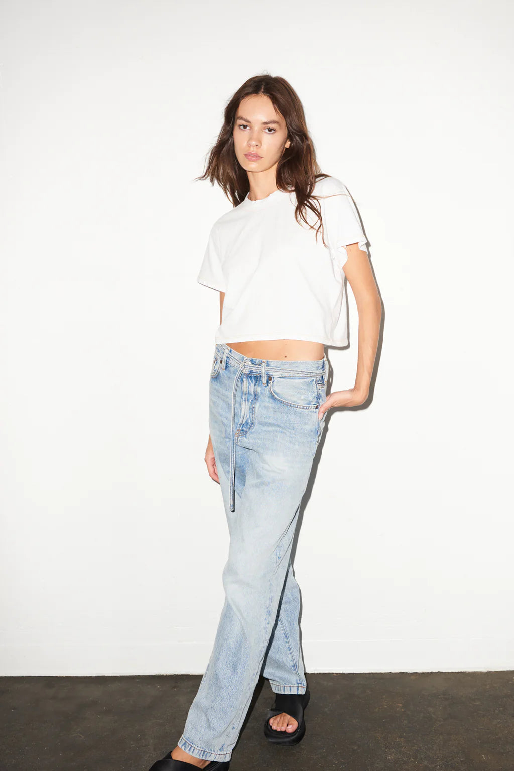 Woman in a white cotton tee and light blue jeans stands by a plain white wall, one hand resting in her pocket.